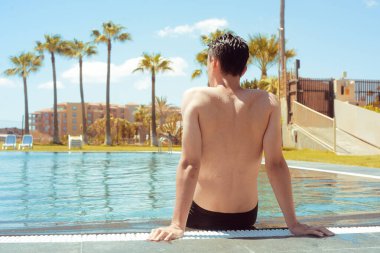 Bir adam ayakları suyun içinde havuz kenarında oturuyor. Güneşli bir gün, bir erkek dinlenmeyi sever. Seyahat et. Bir adam palmiye ağaçları ve bir evin arka planında oturuyor. Otelde havuzlu bir adam..