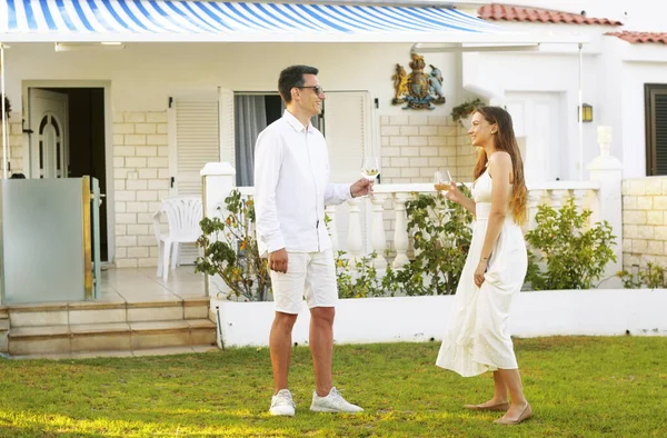 A couple drinks white wine on the lawn near their home on the island. On a trip, the couple rented a house, relax in the sun, the couple laughs and drinks wine. A young couple celebrates a purchase at home. Happiness couples.