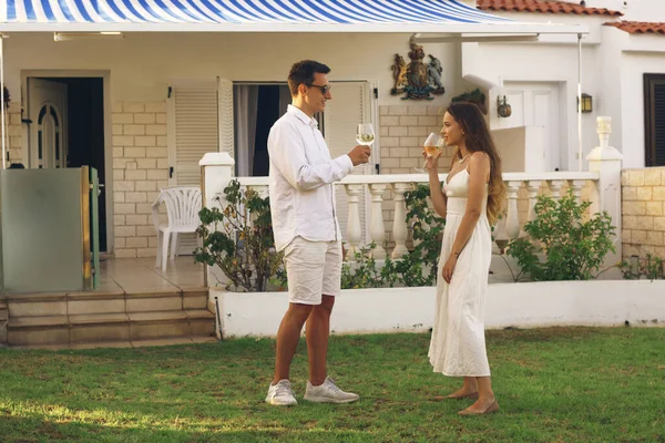 A couple drinks white wine on the lawn near their home on the island. On a trip, the couple rented a house, relax in the sun, the couple laughs and drinks wine. A young couple celebrates a purchase at home. Happiness couples.