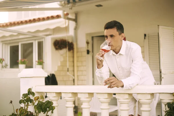 A man stands on a terrace and smokes a cigar, drinks red / pink wine. A rich man is resting in a house on the ocean. Traveling alone, the man thought. A man in white on vacation.