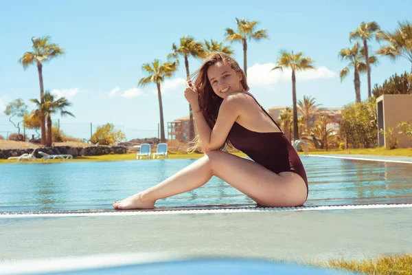 华丽的金发姑娘度假时坐在游泳池边 一个穿着红色泳衣的长发女孩坐在棕榈树的背景上 阳光灿烂的日子 女孩旅行 日光浴 夏天的心情 — 图库照片