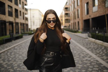 Göz kamaştırıcı kız şehirde dolaşıyor. Esmer, uzun saçlı, siyah elbiseli ve derili. Kız, moda fotoğrafı, sokak modası. Model Avrupa 'yı dolaşıyor, seyahat ediyor. Moda gözlüklü kız..