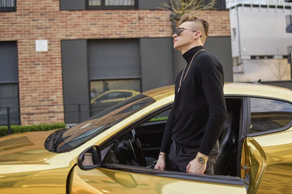 The typical guy is rich, rich parents. Rapper guy in gold chains, gold watch, on a golden car. Teen gets out of his car. A guy with glasses is looking at the camera.