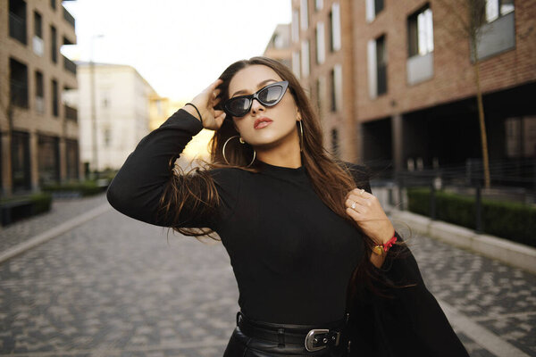 Gorgeous girl walks about the city. Brunette with long hair in a black dress and leather. Girl, fashion photo, street fashion. The model walks around the city of Europe, travels. Girl in fashionable glasses.
