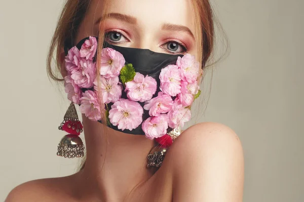 Çiçek maskeli bir kızın moda fotoğrafı. Nefes alamadığımız bahar. Virüs, salgın hastalık, koronavirüs, maskeli manken, taze pembe çiçeklerden oluşan bir maskeli güzellik modeli. Narin pembe makyajlı kız..