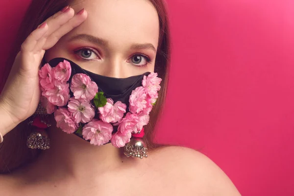 Bahar kızı, çiçek maskeli pembe arka planda bir mankenin portresi. Kız çiçekleri soluyor. Maskeli güzel pembe makyajlı pembe arka planda bir kız. Moda fotoğraf salgını, koronavirüs, karantina.