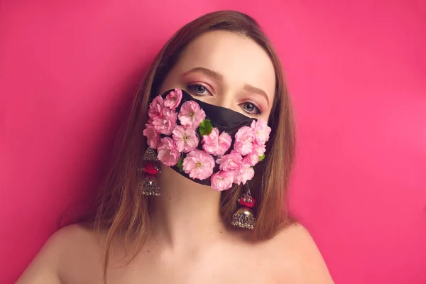 Bahar kızı, çiçek maskeli pembe arka planda bir mankenin portresi. Kız çiçekleri soluyor. Maskeli güzel pembe makyajlı pembe arka planda bir kız. Moda fotoğraf salgını, koronavirüs, karantina.