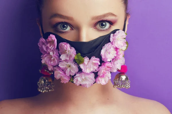 Maske takmış güzel bir bahar kızı. Nefes alamadığımız bahar, çiçeklerin kokusu. Mor arka planda çiçek ve çiçek tomurcukları maskesi takmış bir kız portalı. Koronavirüs, maskeler, salgın hastalıklar.