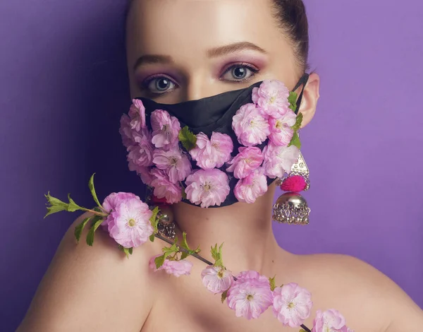 Maske takmış güzel bir bahar kızı. Nefes alamadığımız bahar, çiçeklerin kokusu. Mor arka planda çiçek ve çiçek tomurcukları maskesi takmış bir kız portalı. Koronavirüs, maskeler, salgın hastalıklar.