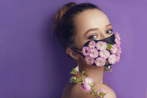 Maske takmış güzel bir bahar kızı. Nefes alamadığımız bahar, çiçeklerin kokusu. Mor arka planda çiçek ve çiçek tomurcukları maskesi takmış bir kız portalı. Koronavirüs, maskeler, salgın hastalıklar.