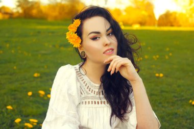 Muhteşem esmer, çimenlerin üzerinde beyaz bir elbiseyle oturuyor ve elinde karahindibalar, sarı karahindiba çiçekleri var. Çiçekli bir tarlanın arka planında şık sarı renkli parlak makyajlı bir kızın portresi. Kızın artık alerjisi yok..