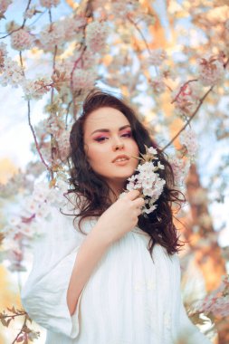 Çiçekli inanılmaz hassas bir kız, pembe çiçekli bir ağacın arka planında, pembe makyajlı bir kızın şık bir fotoğrafı. Baharda esmer, uzun saçlı. Burnu delinmiş bir kız. Güzellik portresi, alerji