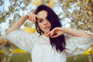 Beyaz çiçekli bir ağacın arka planında, çiçekli, beyaz boho elbiseli bir manken gibi inanılmaz hassas bir kız. Baharda esmer, uzun saçlı. Burnu delinmiş bir kız. Güzellik portresi, alerji.