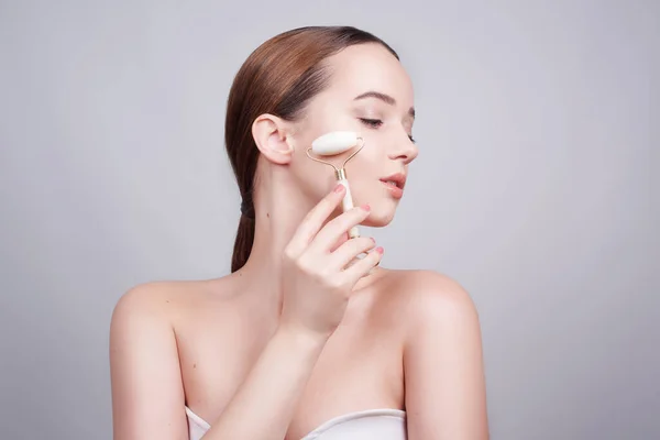 Portrait photo of a young woman looking relaxed use while using a natural white quartz face roller. Beauty face care. Woman doing face massage with jade facial rollers for spa skin care treatment at home.