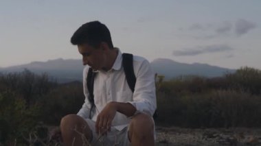 Sırt çantalı bir gezgin dağlarda beyaz bir adada bir kaktüsün yanında oturuyor ve ona dokunuyor..