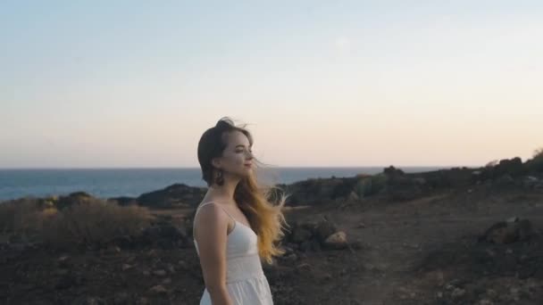 少女は日没時に夏の島を歩く 長い髪をした柔らかい女の子が山の中の白いドレスを着て歩いている 海を背景にしたモデル 穏やかな — ストック動画