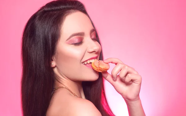 Girl eats macaroon, model on a pink background eats a sweet, sugar diet. Brunette with pink make-up, addicted to food. Macaroon bites, cookies