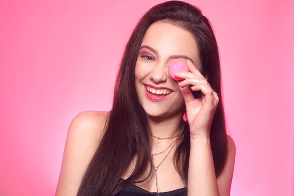 Girl eats macaroon, model on a pink background eats a sweet, sugar diet. Brunette with pink make-up, addicted to food. Smiles