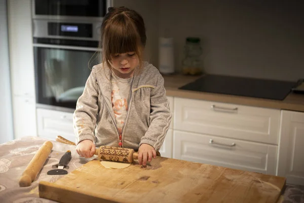 Child girl, 3 years old, in white kitchen, flattening pizza dough with a small rolling pin on a wooden board