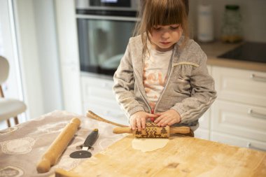 Çocuk, 3 yaşında, beyaz mutfakta, pizza hamurunu tahta bir tahtada merdaneyle düzleştiriyor. Anaokulu öğrencileri için kilitleme etkinliği fikri.