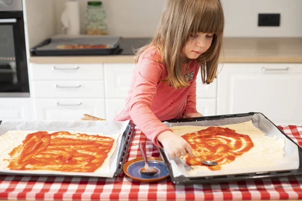 Pişirmeden önce mutfakta çiğ pizzaya domates sosu döken bir kız çocuğu. Çocuklar için tecrit aktivite fikri. Ön görünüm.