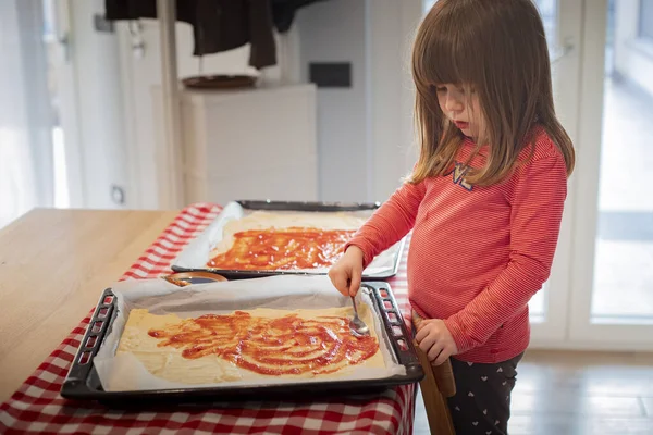 Çocuk kız sandalyeye çıkıp pişirmeden önce çiğ pizzaya domates sosu sürüyor. Çocuklar için tecrit aktivite fikri. Yan görünüm.