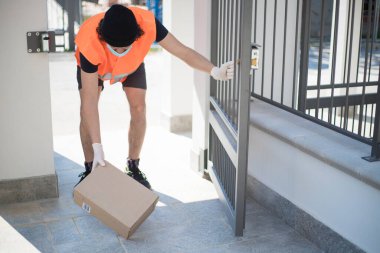 Teslimat şoförü açık kapının hemen arkasında bir paket kutusu bırakıyor. Kapatma sırasında eldiven ve yüz maskesi takıyor. Dışarıdan paket getiren bir adam var..