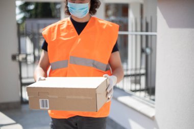 Kapıda paket kutusu olan genç bir teslimatçı var. Kapalı hava salgını sırasında eldiven ve maske takıyor. Kafkas sürücüsü açık hava şartlarında bir paket teslim ediyor..