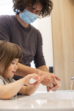 Salgını önlemek için yüz maskesi takan beyaz bir baba covid-19 salgını sırasında küçük kıza tuvalette nasıl el yıkaması gerektiğini öğretiyor. Baba ve küçük kız lavaboda el yıkıyorlar. Dikey çekim.