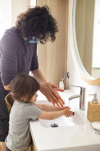 Caucasian father wearing face mask to avoid contagion teaching little child girl how to wash hands in bathroom during covid-19 pandemic lockdown. Dad and baby daughter washing hands at sink. Vertical shot.