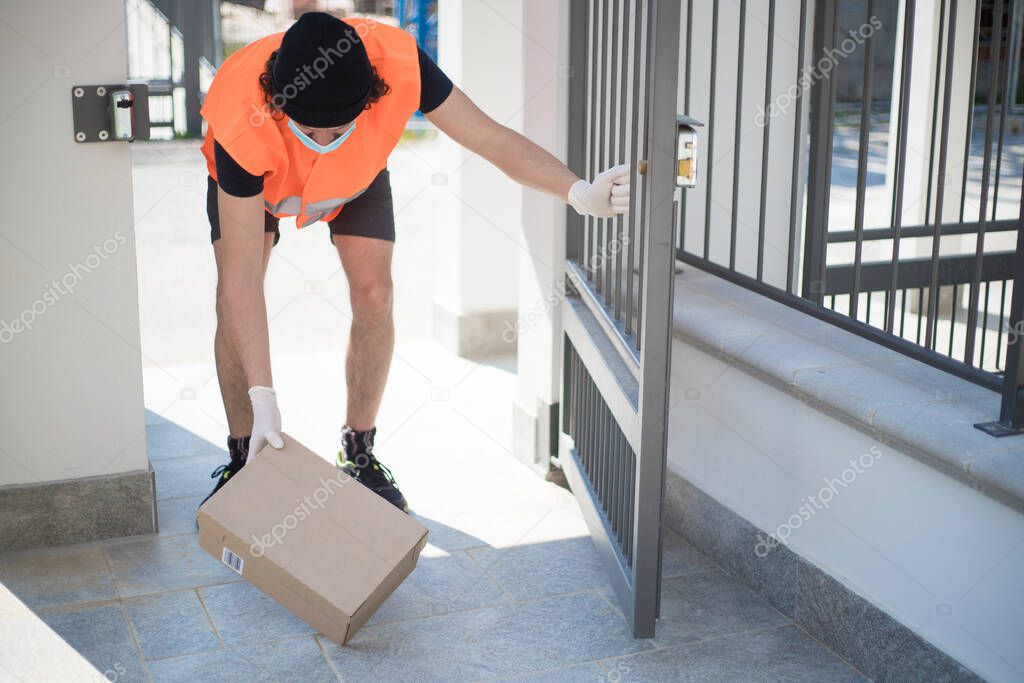 Delivery driver leaving a parcel box on the floor just beyond the open gate, wearing gloves and face mask during covid-19 pandemic lockdown. Man delivering a package inside the front door in outdoor context.