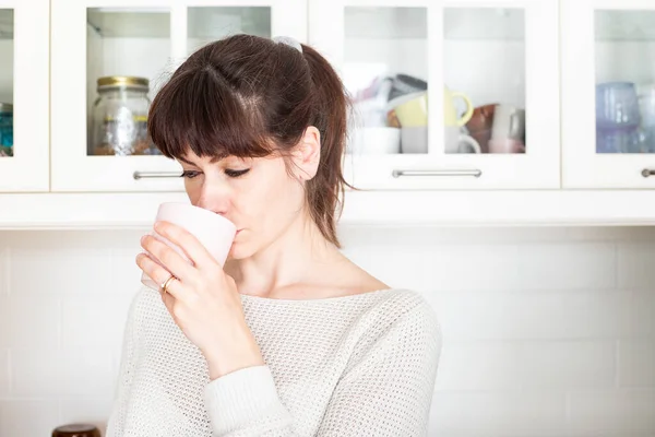 Pretty caucasian woman, with bangs and ponytail, drinking a cup of coffee in the morning in a white, bright lit, kitchen. Looking away.