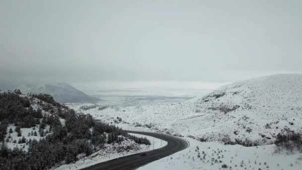 En drönare reser sig ovanför en snöig skog. I fjärran skyms ett berg av dimma. Bilar rör sig längs vägen i ett svårt område med en skarp sväng, en serpentin. — Stockvideo