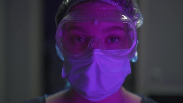 若い医者の肖像少女は眼鏡 医療用マスク 科学者の頭の上のキャップを修正します 紫色の照明のあるオフィスでコロナウイルスワクチンを作成する — ストック動画