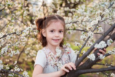 Güzel bir kızın bahar portresi. Kahverengi gözlü şık bir kız. Beyaz çiçekli küçük bir kızın portresi. Beyaz çiçek açan ağaç ve mutlu bebek