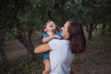 Bebek annesinin ellerinde yüksek sesle gülüyor. Dışarıda sonbahar akşamının aile portresi. Genç anne ve mutlu kız.