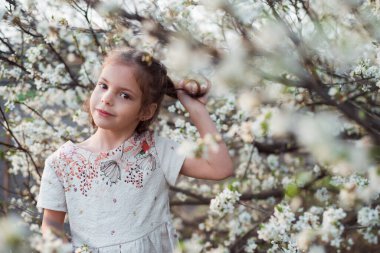 Çiçek açan bir ağacın arka planında bir çocuğun bahar portresi. Kahverengi gözlü sevimli kız elleriyle saçlarını tutuyor.