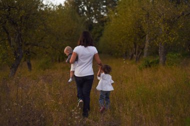 Genç bir anne gün batımından sonra çocuklarıyla birlikte sarı çimlerde yürür. Parkta sonbahar yürüyüşü