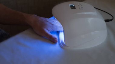 Kadın, beyaz masaya akrilik manikür yaptığı için LED lambanın içine elini sokuyor. Manikür için mavi lamba. Çivilerin üzerindeki laktürün kuruması