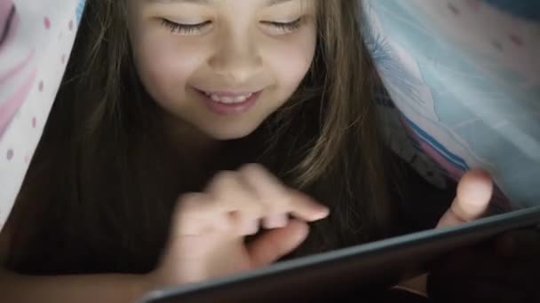Menina de dez anos usa tablet digital sob cobertura, à noite — Vídeo de Stock