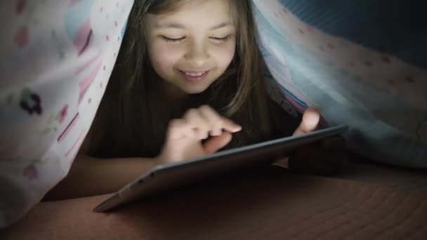 Niña utiliza tableta digital bajo cubierta, por la noche — Vídeos de Stock