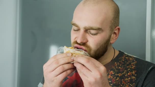 Молодой человек ест вкусный сэндвич, стоит в помещении — стоковое видео
