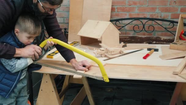 Un petit garçon apprenant à utiliser une scie à main sur un panneau en bois — Video