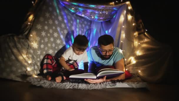 Father and son read inside a tent set up indoors — ストック動画
