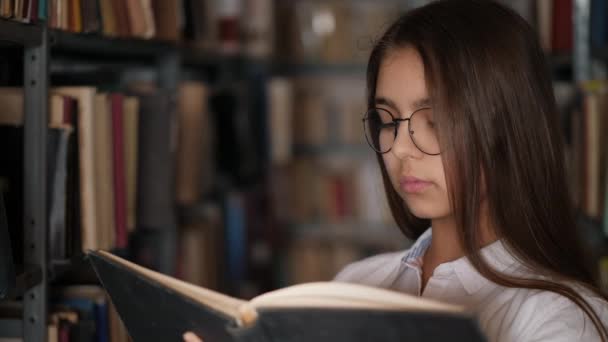 Egy fiatal lány portréja, aki könyvet olvas egy könyvtárban. — Stock videók
