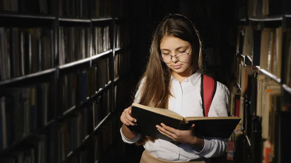 Menina com óculos lendo um livro na biblioteca — Fotografia de Stock
