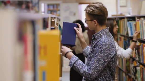 Un joven con gafas elige un libro en una biblioteca — Vídeos de Stock