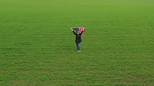 アメリカの国旗が描かれた医療用マスクや眼鏡の女性の空中風景が緑の野原の真ん中に立っている 国家の心理的支援 — ストック動画
