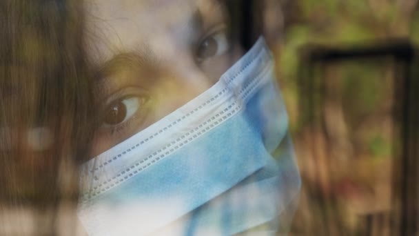 Una adolescente con máscara médica mira por la ventana — Vídeos de Stock