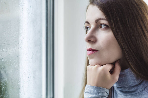 A sad, pensive young woman, in rainy weather, sits at the window. The woman is alone.